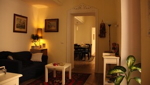 A living room with a dark-colored sofa, a white coffee table, and a red patterned rug. There is a white lamp on a side table, and a potted plant to the right. - Il Conservatorio B&B (Naples)
