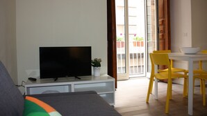 A living room with a flat-screen TV on a white stand, a grey sofa, and a dining area with a white table and yellow chairs. - Il Conservatorio B&B (Naples)