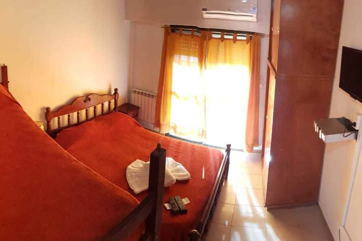 A single bed with a wooden frame and a red bedspread in a room with a window, a wall-mounted TV, and a radiator.