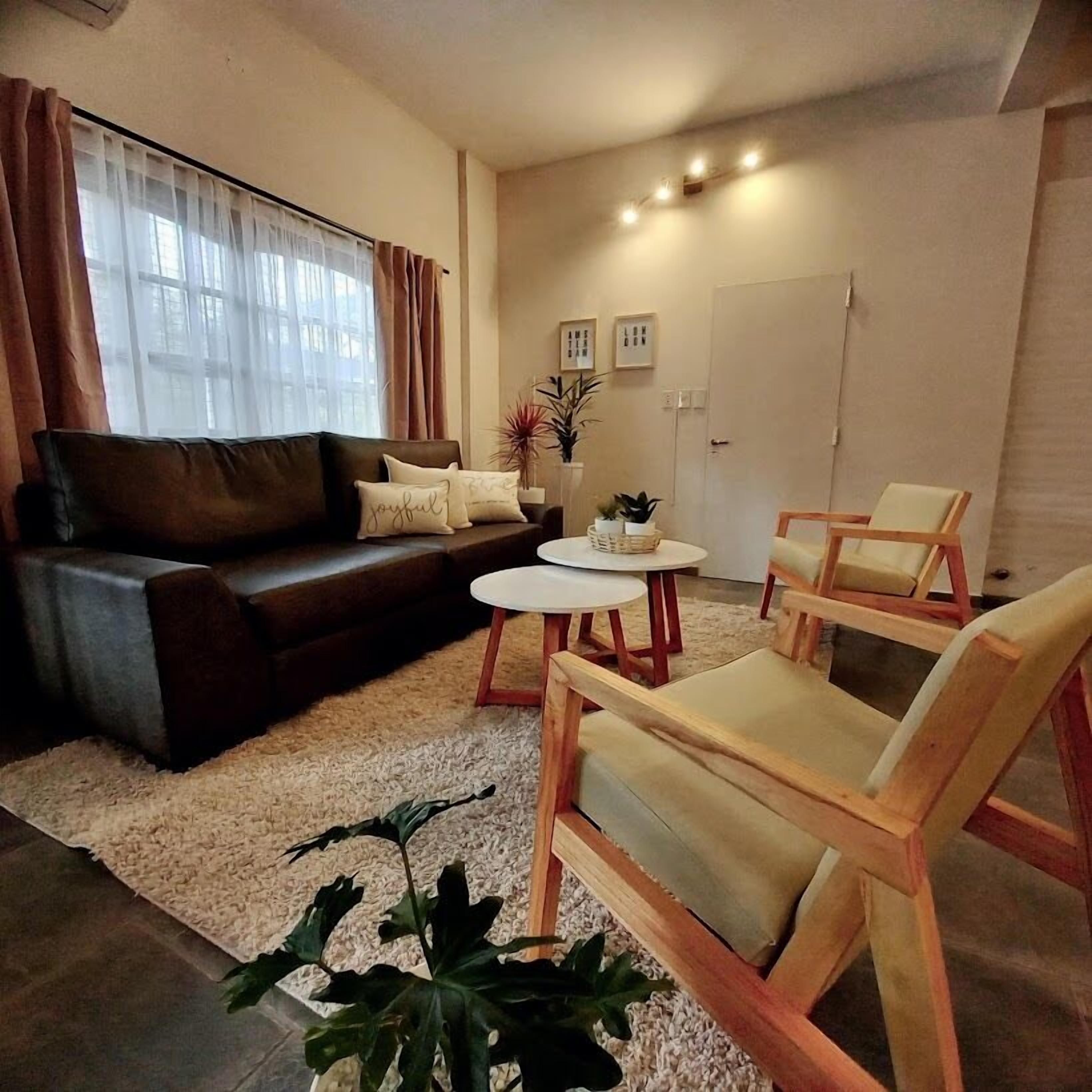 Una sala de estar con un sofá marrón oscuro, dos sillas de madera, una mesita redonda y un ventanal con cortinas de gasa.