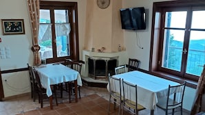 Uma sala de jantar com uma mesa branca, cadeiras e uma lareira. Há um relógio na parede e uma TV instalada acima da lareira.