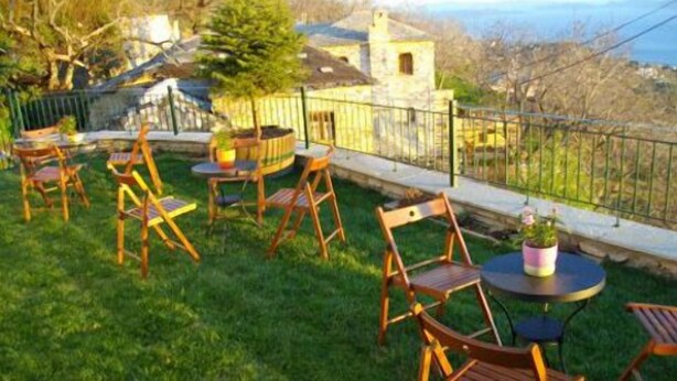 Terraço com cadeiras e mesas de madeira, uma pequena planta em vaso e vista para um edifício de pedra e árvores.