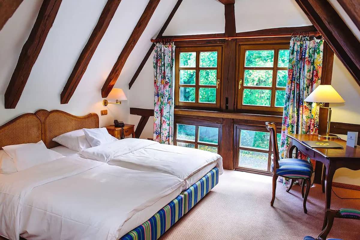 Ein Schlafzimmer mit einem großen Bett, einem Schreibtisch und einem Stuhl. Zwei Fenster mit Vorhängen sind vorhanden.