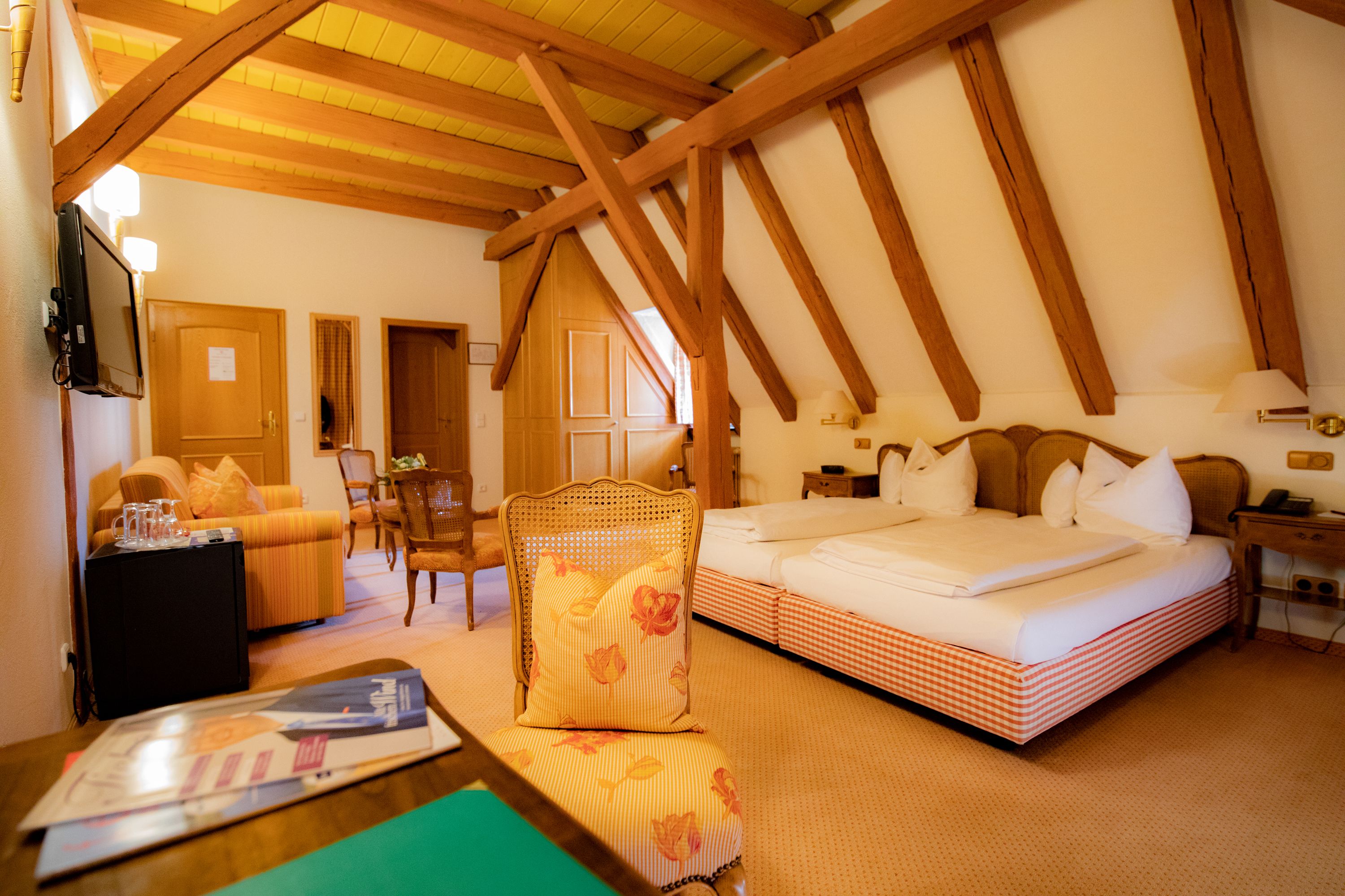 A hotel room with a wooden ceiling, a bed with white bedding, a chair with a floral cushion, a small table with a magazine, and a nightstand with a lamp.