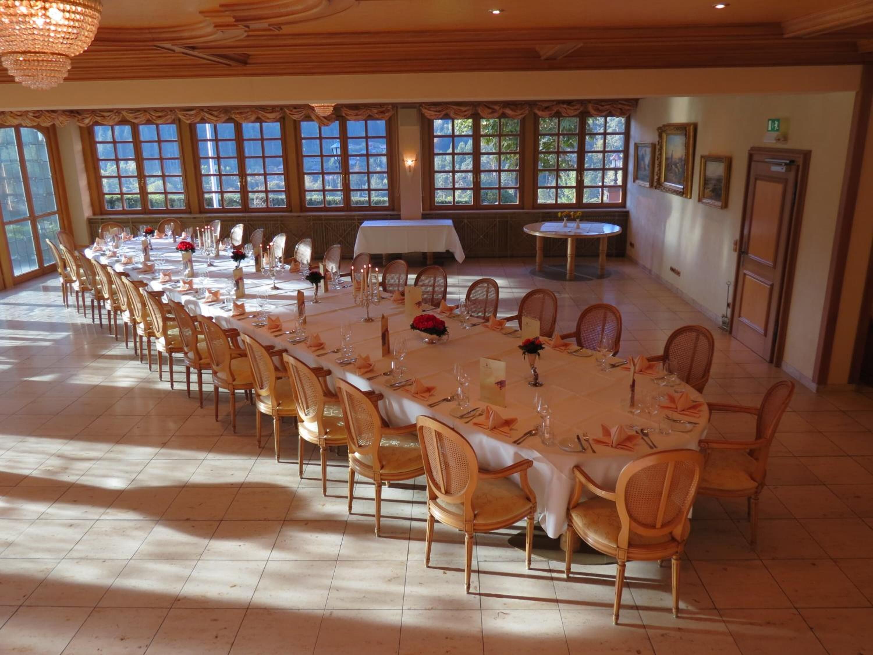 Une salle de banquet avec de longues tables dressées pour le repas, des chaises en bois et de grandes fenêtres ornées de rideaux.