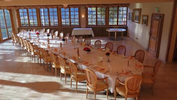 Une salle de banquet avec de longues tables dressées pour le repas, des chaises en bois et de grandes fenêtres ornées de rideaux.