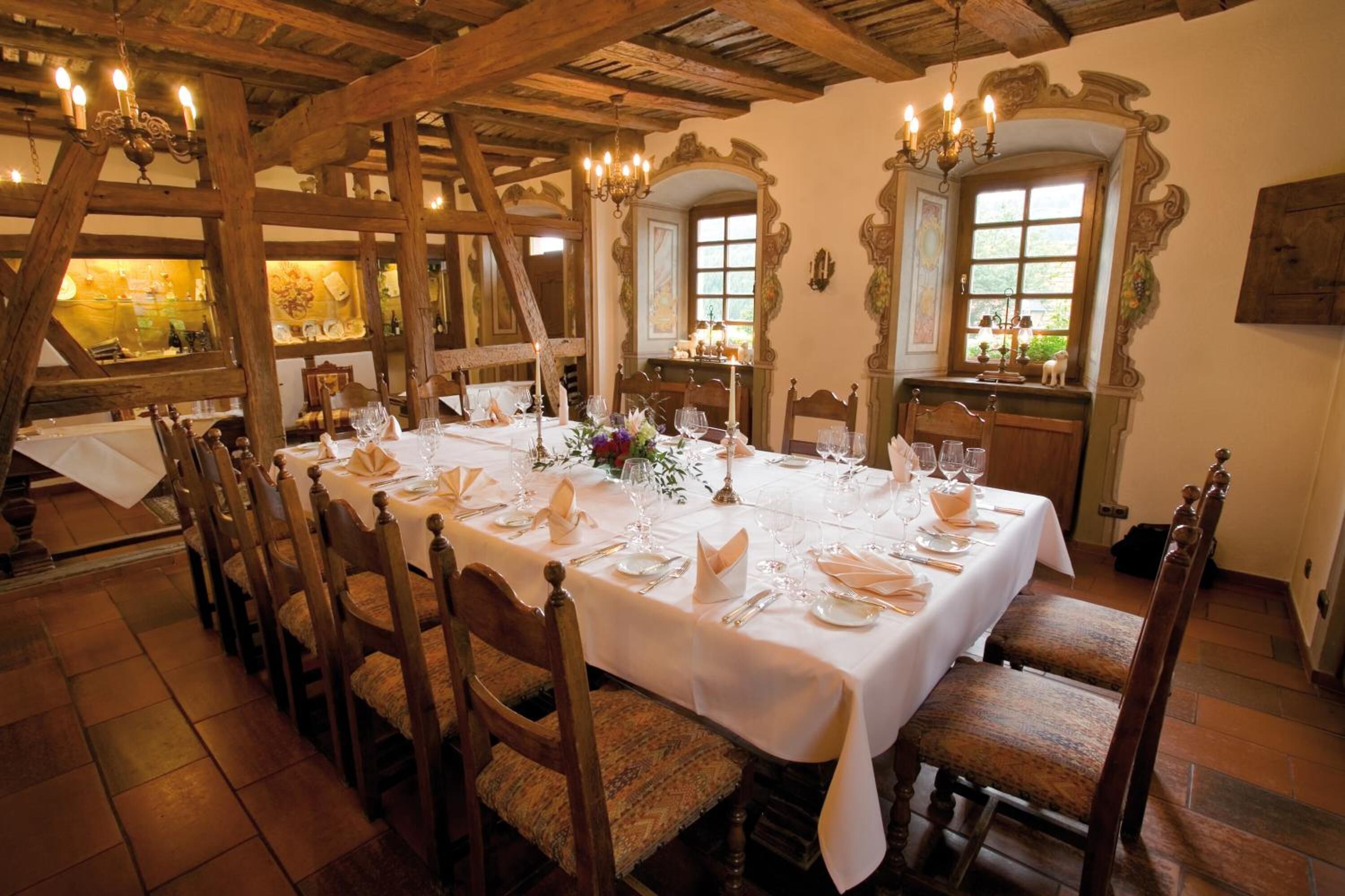 Een eetzaal met een lange tafel gedekt voor een maaltijd, houten stoelen en een rustiek plafond met zichtbare balken.