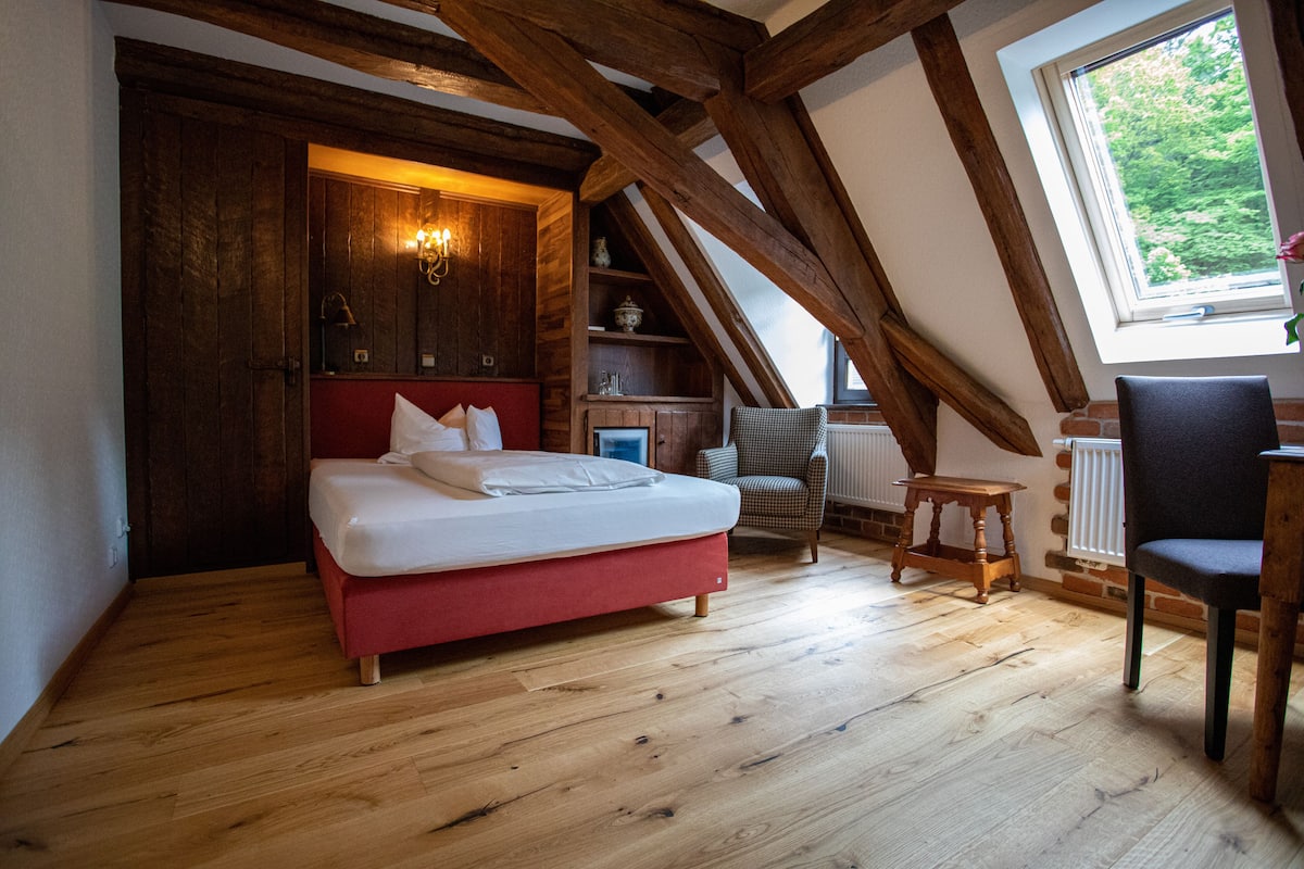 Ein Schlafzimmer mit Holzboden, einem roten Bett, einem Sessel, einem kleinen Tisch und einem Fenster mit Blick ins Grüne.
