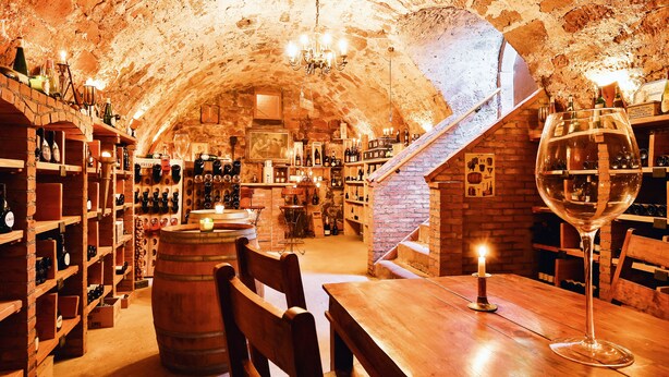 An underground wine cellar with wooden shelves, barrels, and a table with a wine glass. - Hotel Der Schafhof Amorbach (Amorbach)