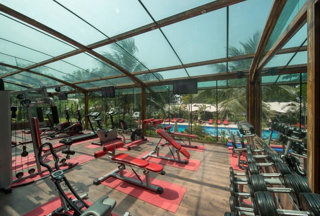 Un gimnasio con diferentes equipos de ejercicio, una piscina y vistas tropicales a través de amplios ventanales.
