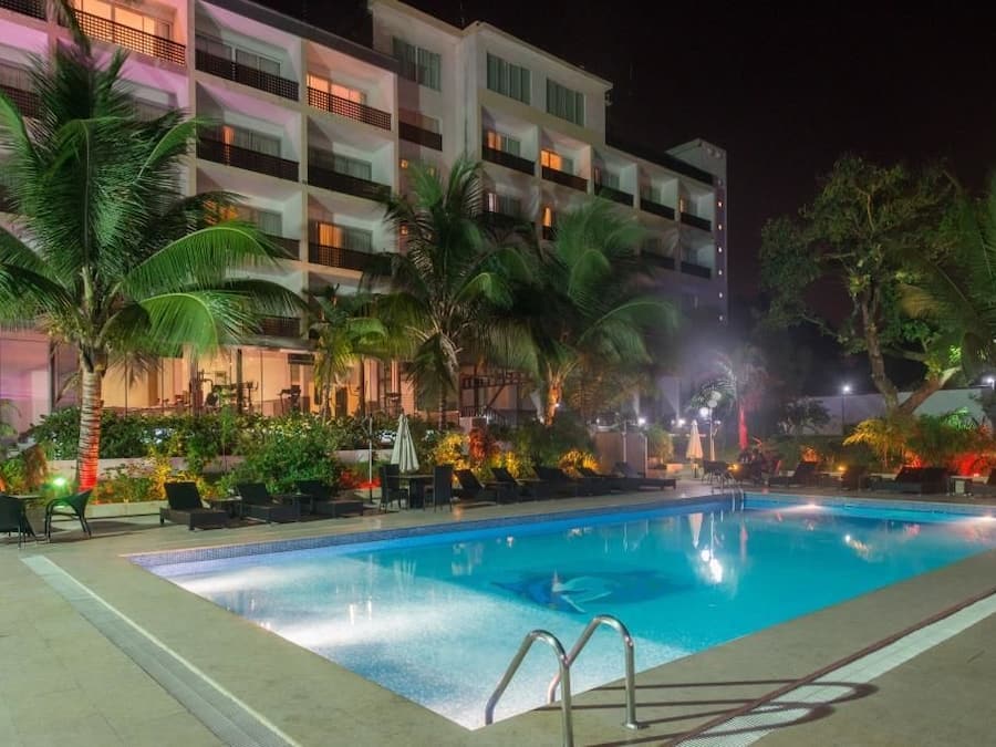 Une piscine entourée de chaises longues et de palmiers, le tout bordé d’un immeuble à plusieurs étages doté de balcons.