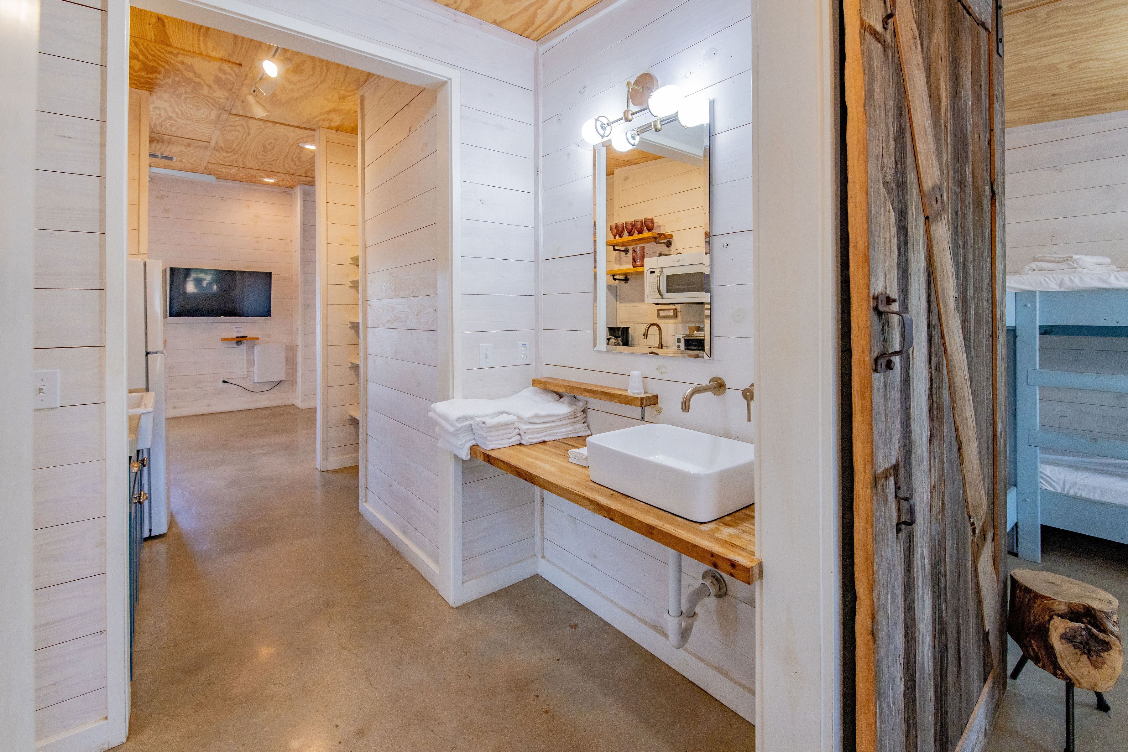 A modern bathroom with white wooden walls, a wooden vanity, a large mirror with lights, a sink, and a television in the background.