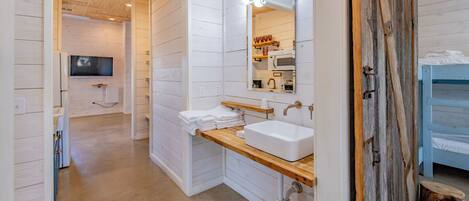 A modern bathroom with white wooden walls, a wooden vanity, a large mirror with lights, a sink, and a television in the background.