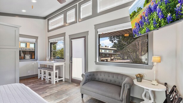 A modern room with a bed, a grey sofa, a small table with a lamp, and a large window with a view of flowers. - 7A Ranch (Wimberley)