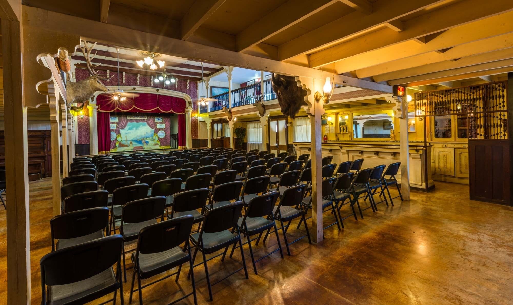 A theater with rows of chairs, a stage with a red curtain, and a balcony.