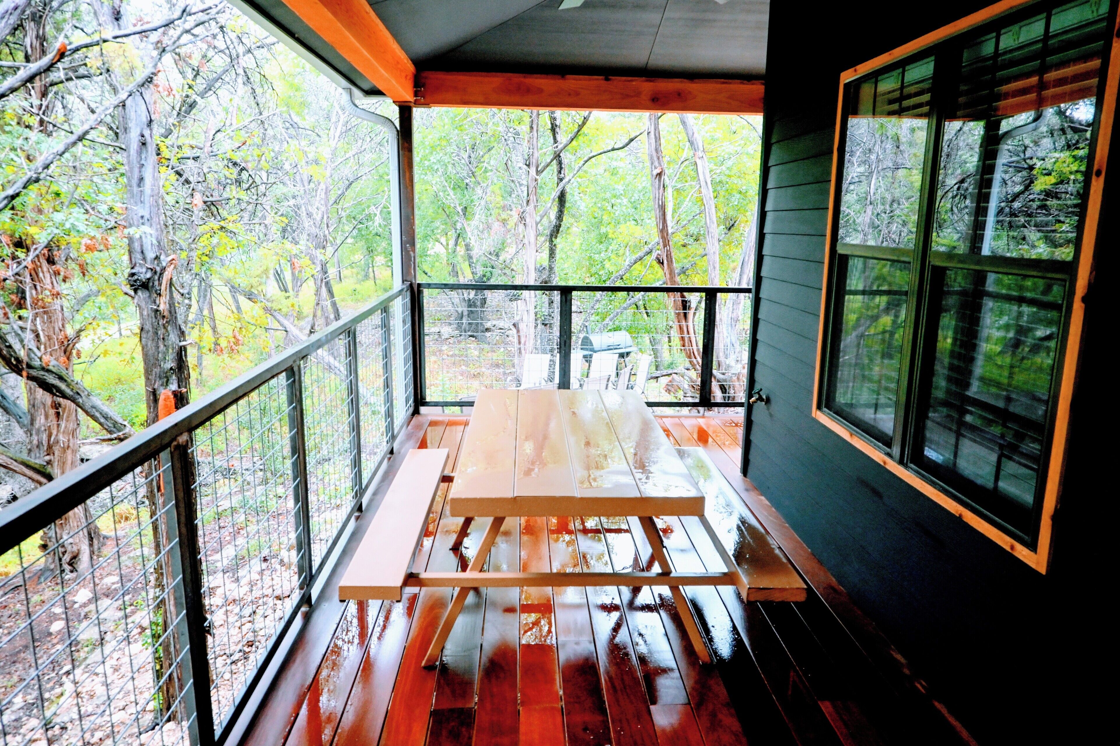 A covered outdoor seating area with a picnic table, surrounded by a wooden deck and metal railings, overlooking a forested area.