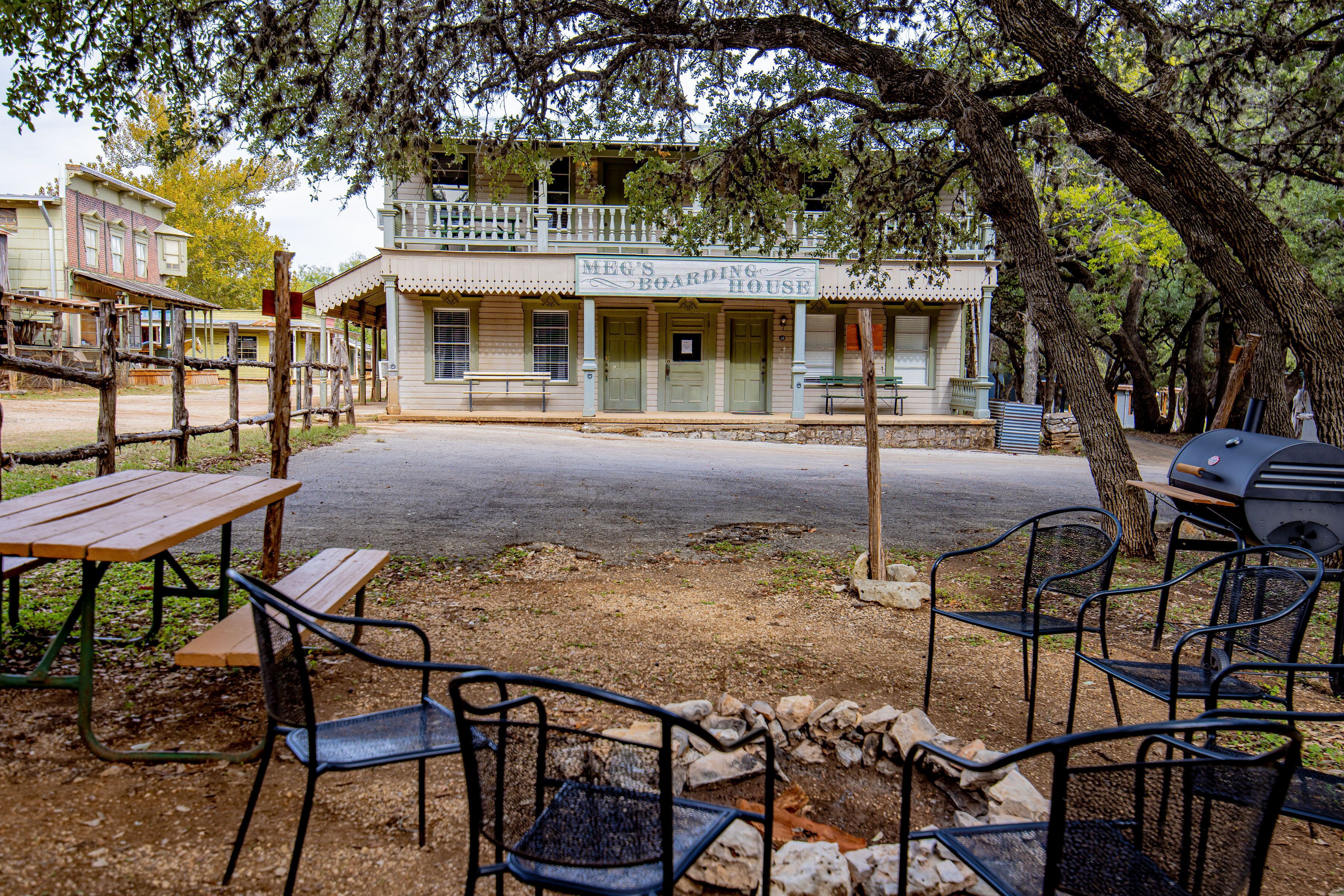 A rustic building with a sign reading "MEG'S BOARDING HOUSE". There are picnic tables and chairs outside, and a barbecue grill is visible.
