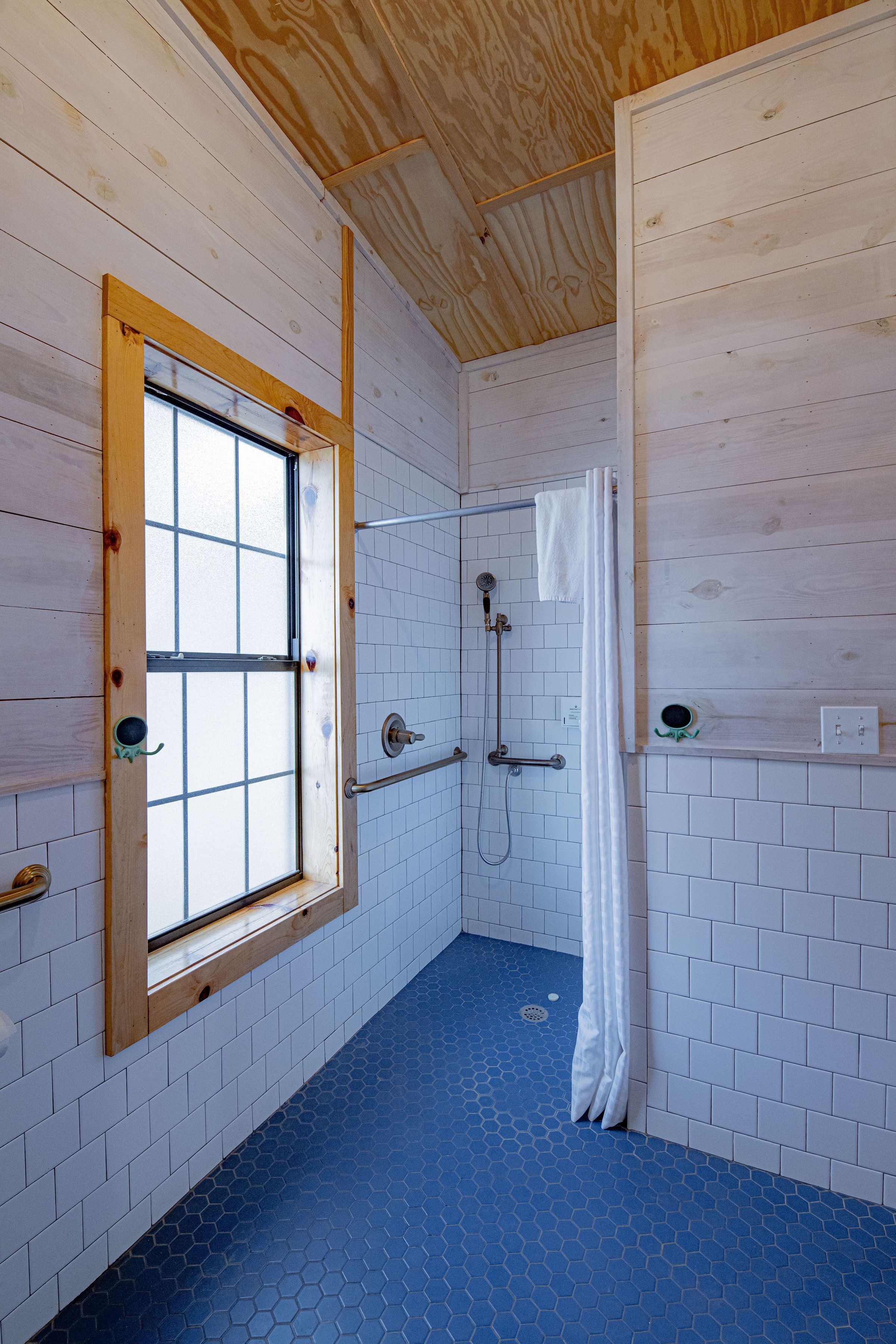 A bathroom with a wooden ceiling, a window with a wooden frame, a shower area with a towel hanging, and a blue hexagonal floor tile pattern.
