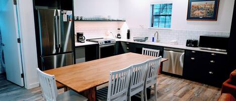 Una cocina moderna con una mesa de comedor de madera, sillas blancas, electrodomésticos de acero inoxidable y una ventana con persianas.