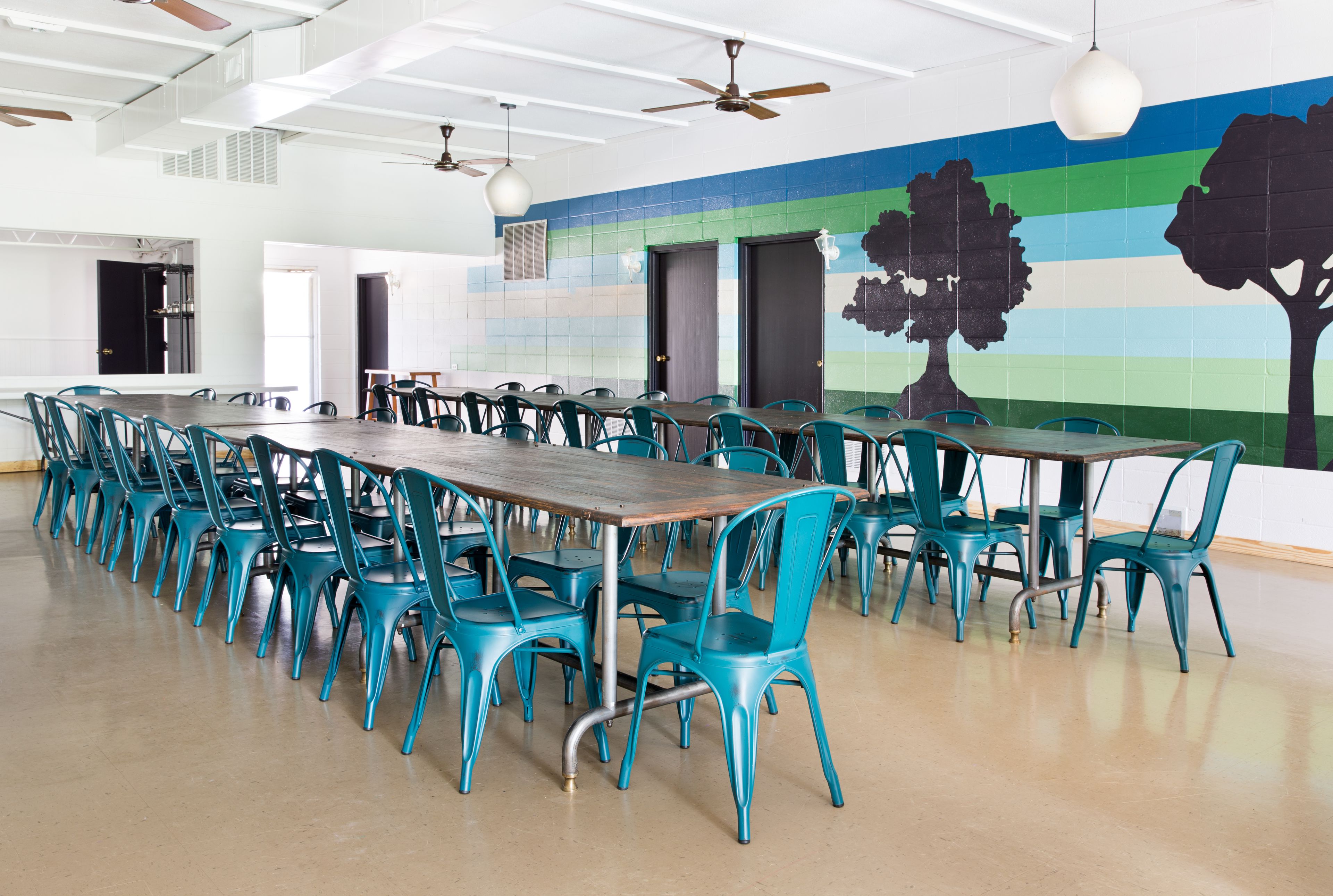 Un comedor amplio con mesas largas y sillas color turquesa, con un mural de árboles en la pared.