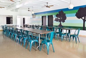 A spacious dining area with long tables and teal chairs, featuring a mural of trees on the wall.