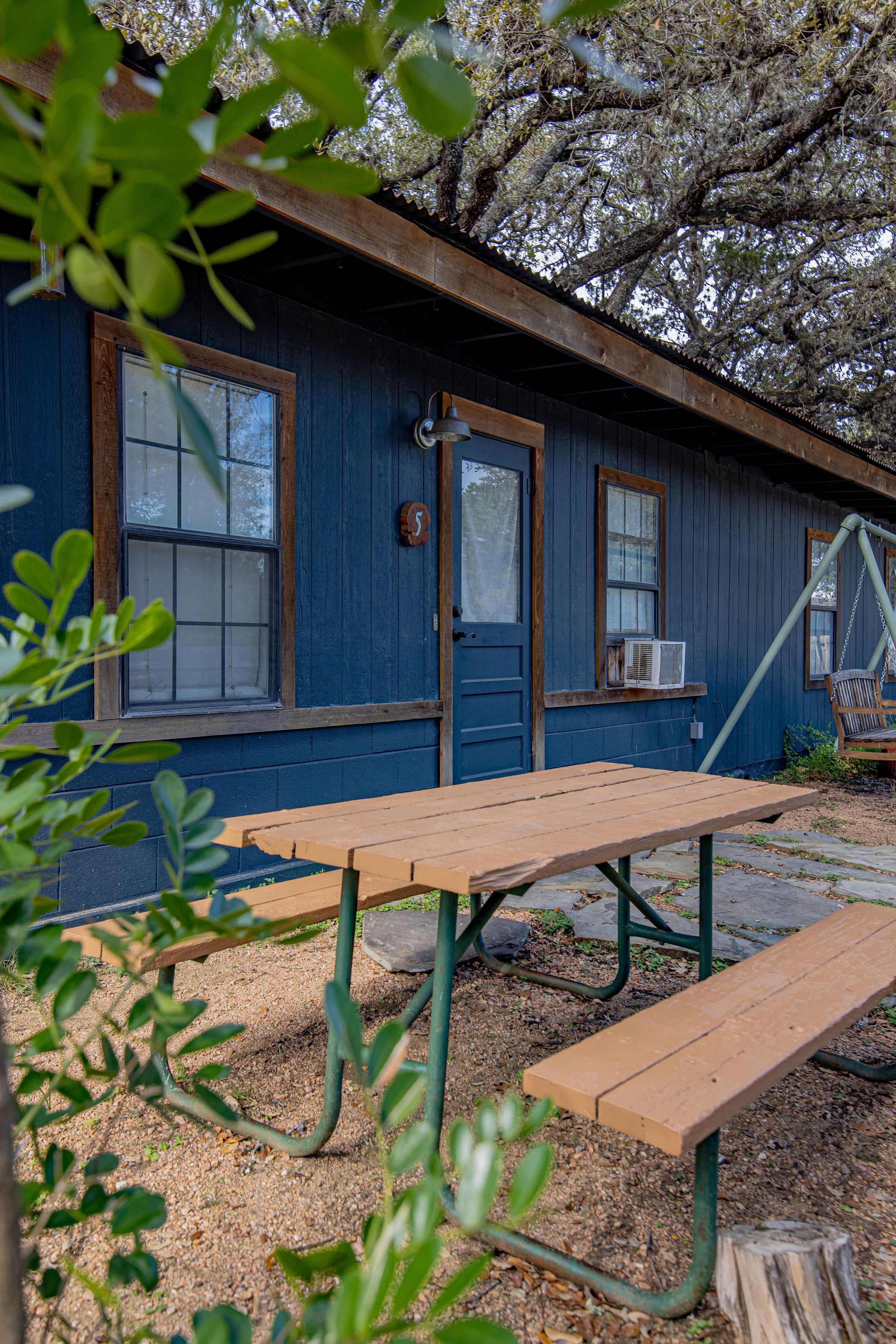 A wooden picnic table and benches in front of a blue building with windows and a door.