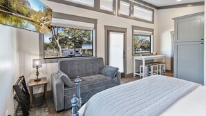 A bedroom with a bed, a grey sofa, a desk with a sink, and a large window with blinds. - 7A Ranch (Wimberley)