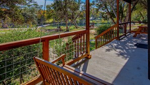 A covered outdoor area with a swing, wooden railings, and a view of a park with trees and a picnic table. - 7A Ranch (Wimberley)