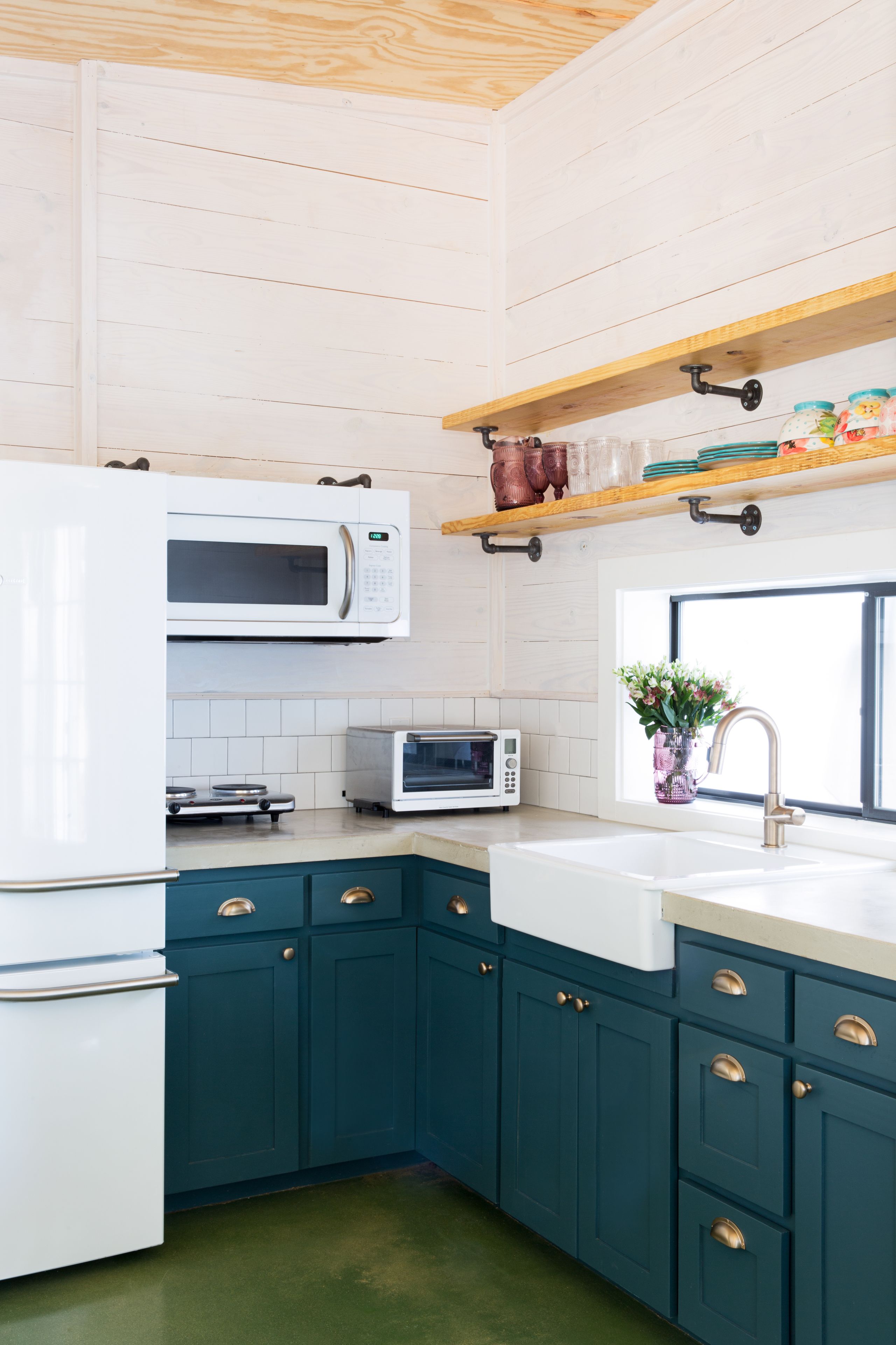 Une cuisine avec des armoires de couleur turquoise, des appareils électroménagers blancs et une étagère en bois au-dessus de l’évier.