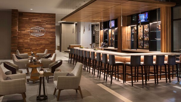 A modern bar area with a long counter, bar stools, and a wooden accent wall with a logo. - Embassy Suites by Hilton Amarillo Downtown (Amarillo)