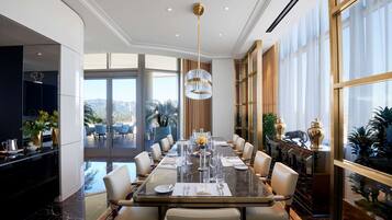 Une salle à manger spacieuse avec une longue table dressée pour un repas, entourée de chaises, un lustre au plafond et de grandes fenêtres offrant une vue dégagée.