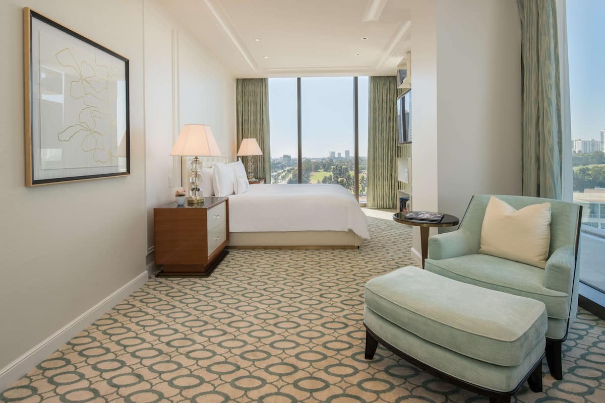 Une chambre d’hôtel avec un grand lit, un fauteuil vert, une moquette à motifs et une vue sur la ville par la fenêtre.
