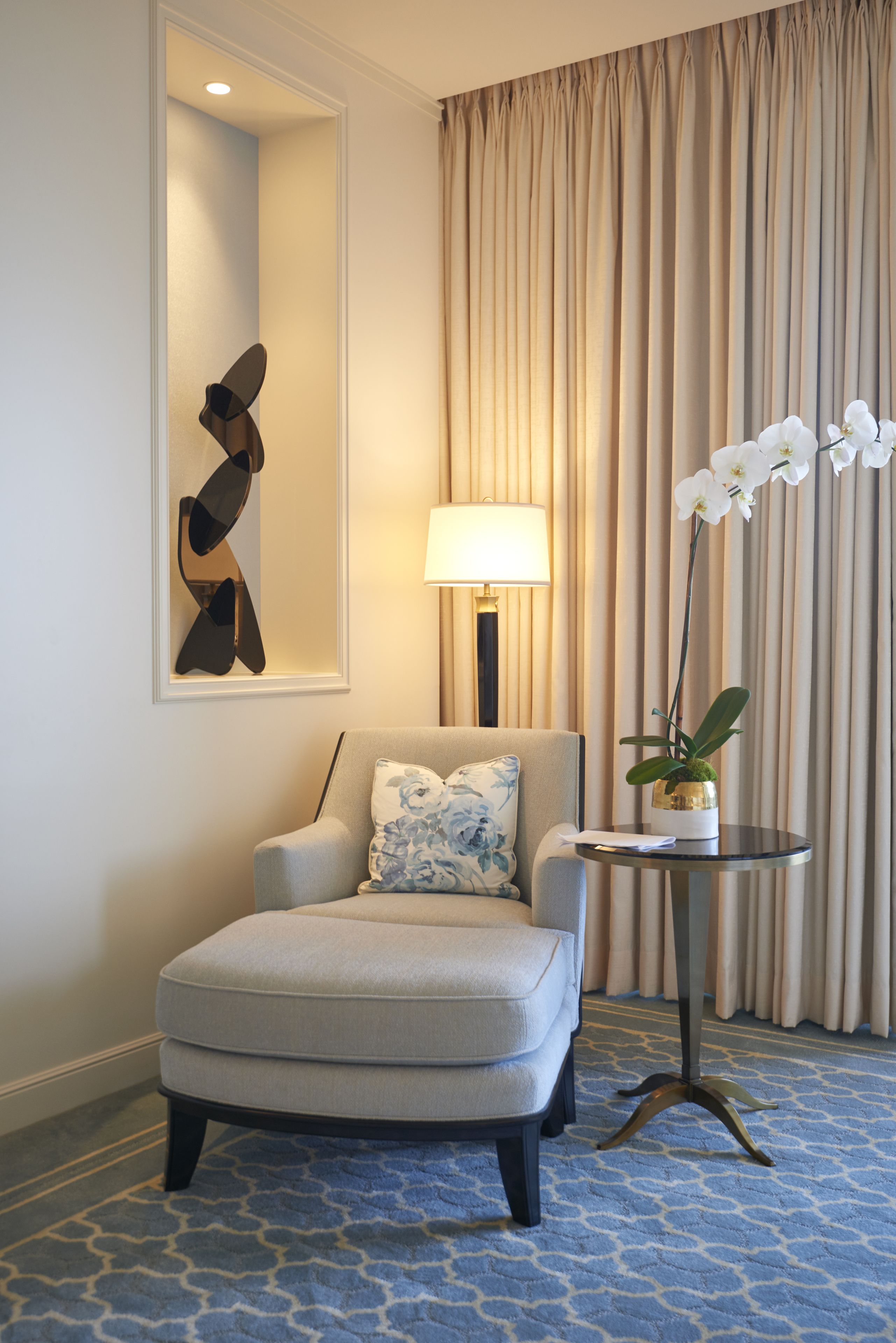 A hotel room with a grey armchair, a round table with a potted orchid, a lamp, and a large mirror on the wall.