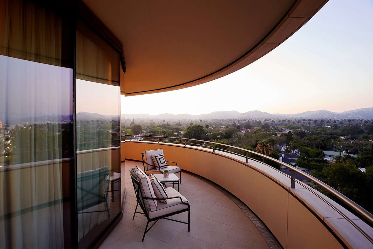 Un balcon doté d’une balustrade incurvée, de deux chaises, d’une petite table et offrant une vue sur un paysage urbain avec des montagnes à l’horizon.
