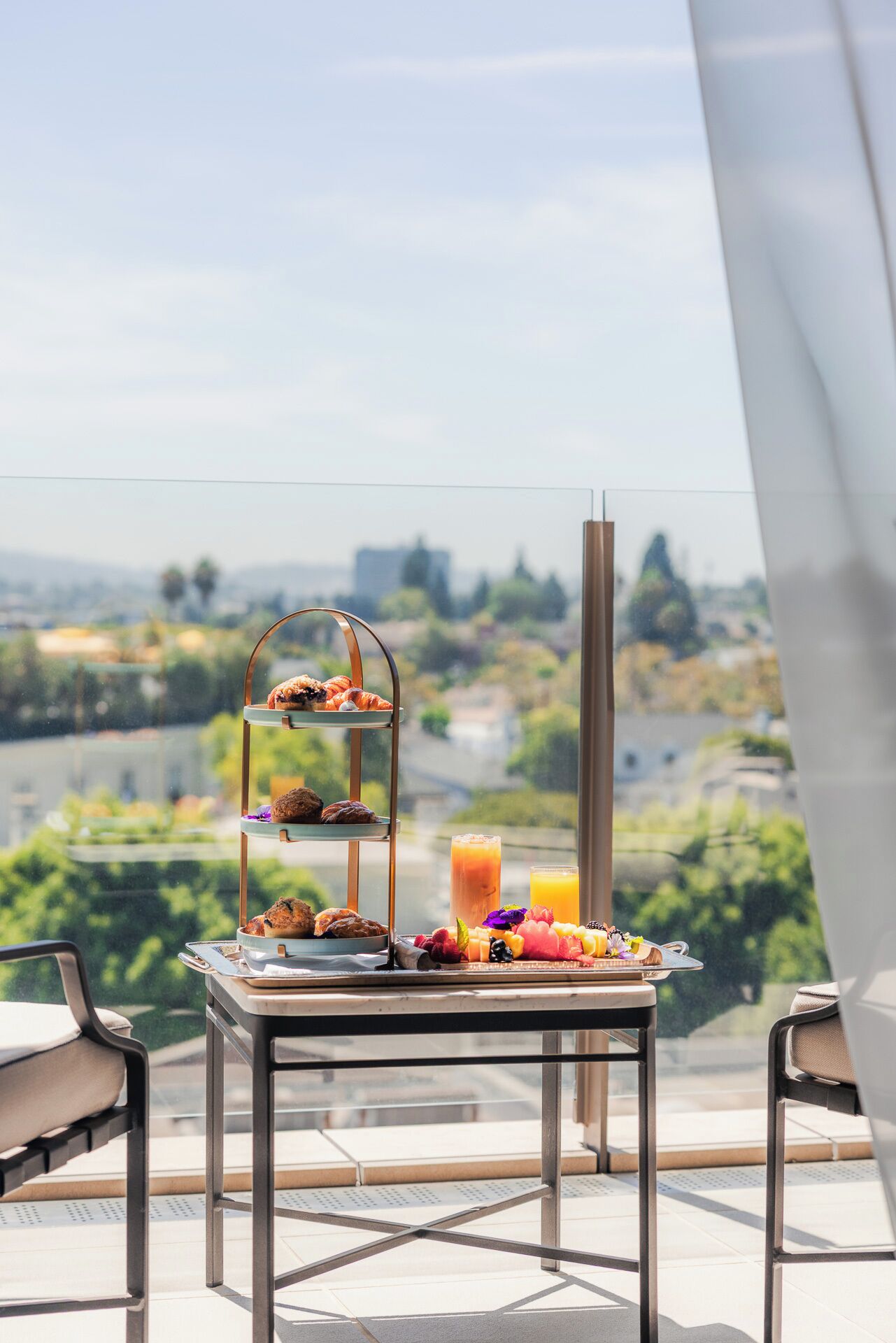 Desayuno en un balcón con vista a la ciudad, incluye un frutero escalonado, un vaso de zumo de naranja y un vaso de café helado.