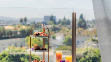 Desayuno en un balcón con vista a la ciudad, incluye un frutero escalonado, un vaso de zumo de naranja y un vaso de café helado.