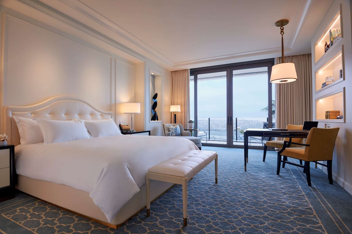 Une chambre d’hôtel avec un grand lit, un bureau avec des chaises, une vue sur la ville et un balcon avec une balustrade.