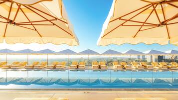 An indoor swimming pool with lounge chairs and umbrellas, with a cityscape view in the background.