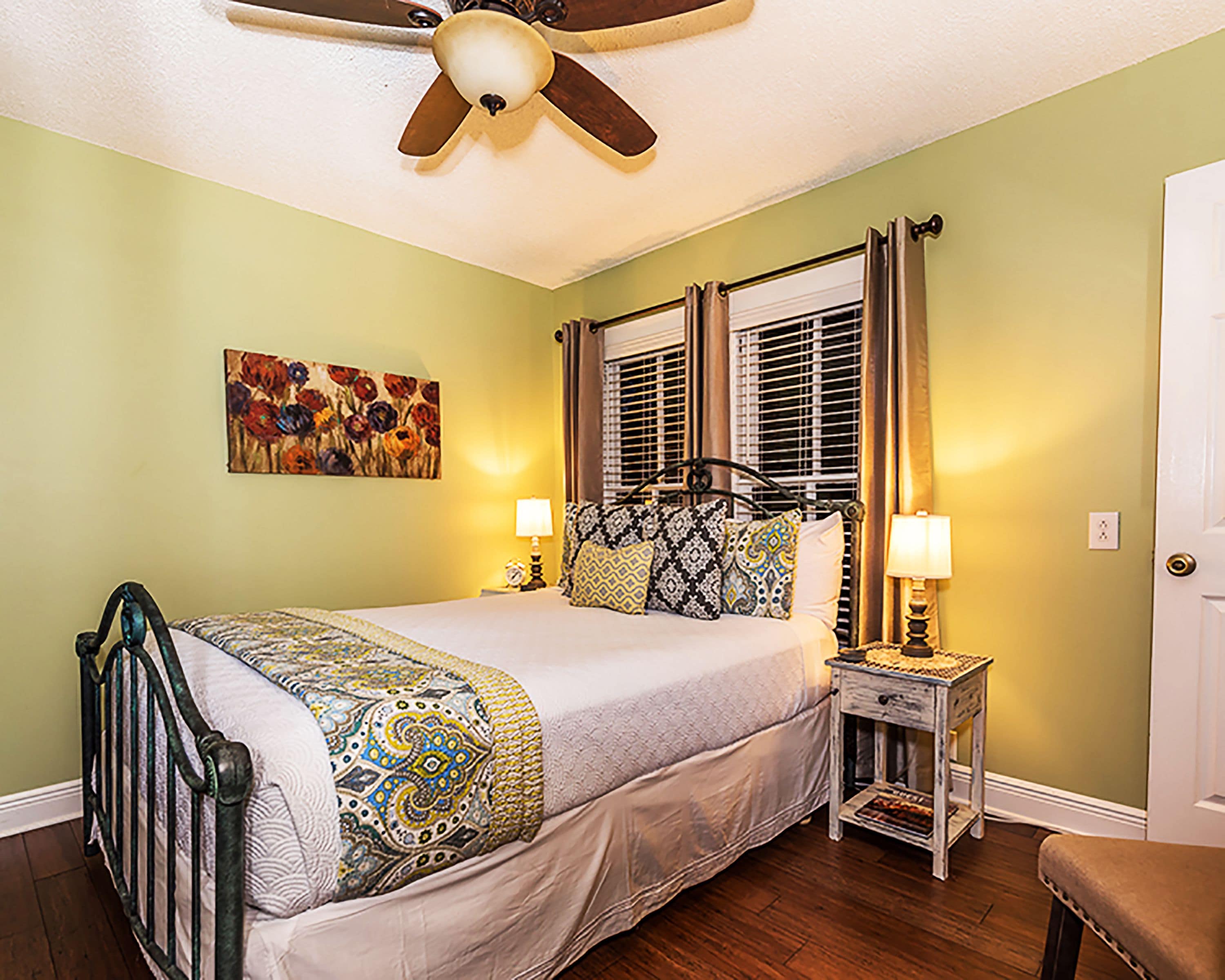 A bedroom with a bed, two bedside tables, a ceiling fan, a window with blinds, and a door.