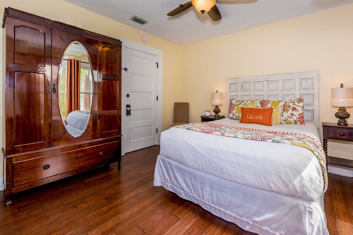 A bedroom with a wooden wardrobe, a bed with a floral headboard, and a nightstand with a lamp.