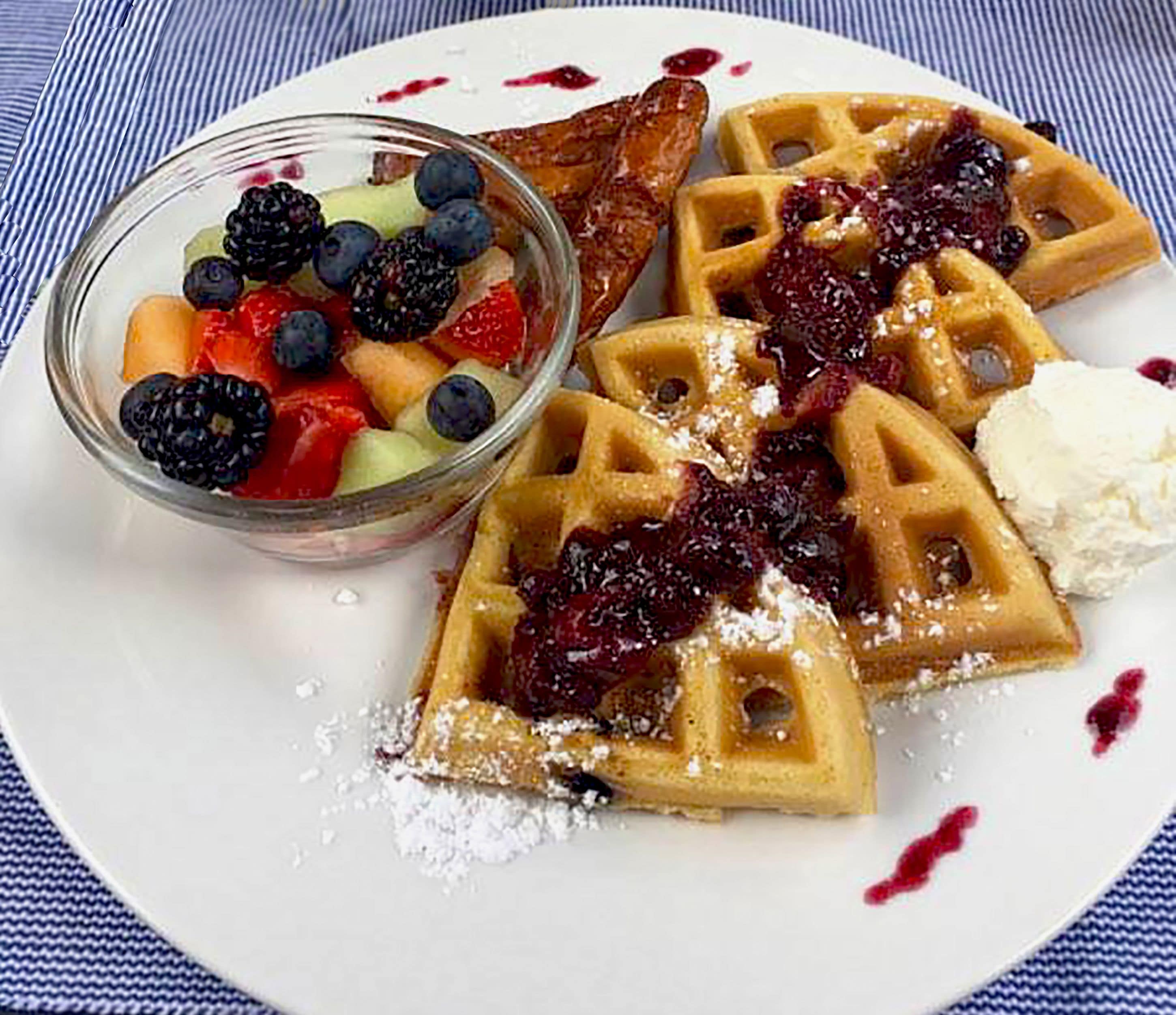 Un piatto con waffle ricoperti di confettura di frutta e zucchero a velo, accompagnato da una ciotola di frutti di bosco e un cono di gelato.