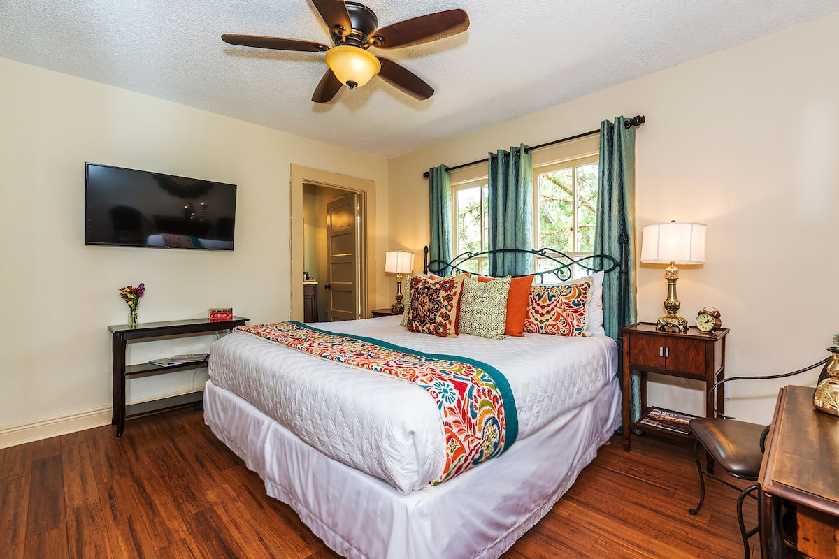 A bedroom with a bed, a desk, a chair, a ceiling fan, a TV, and a window with curtains.