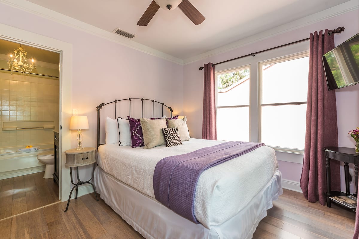 A bedroom with a bed, a nightstand, a lamp, a window with curtains, and a TV on the wall.