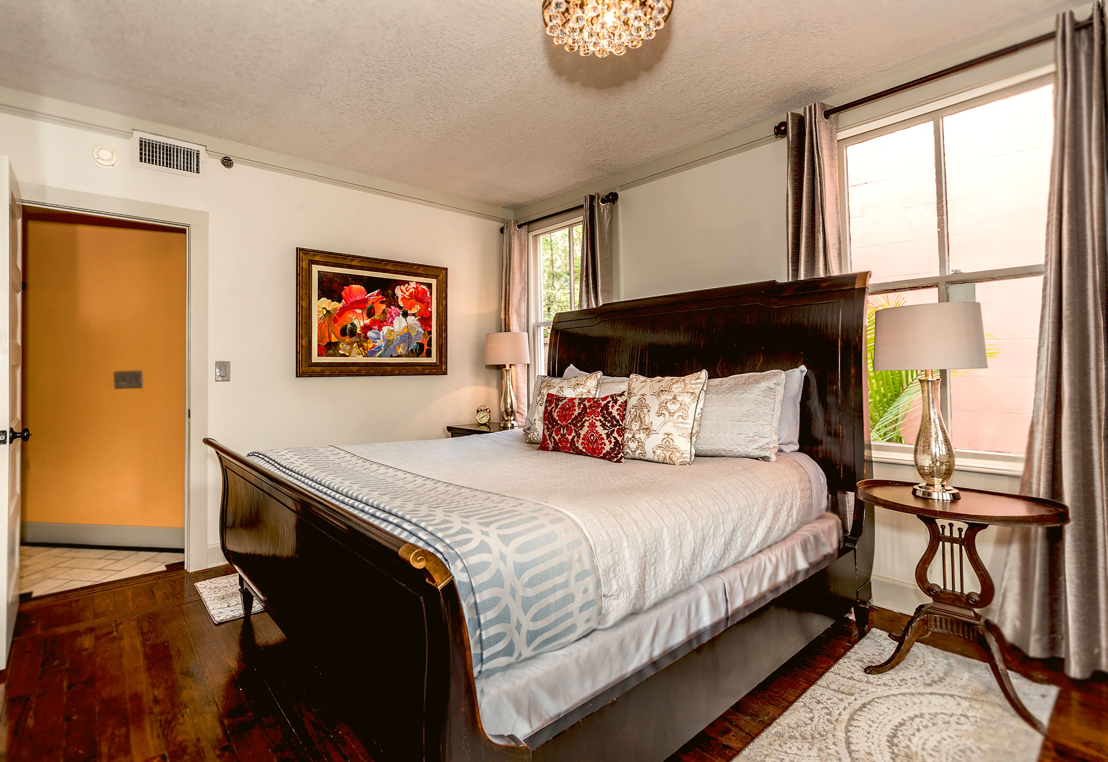 A bedroom with a large bed, a wooden headboard, a nightstand with a lamp, a framed picture on the wall, and a window with curtains.