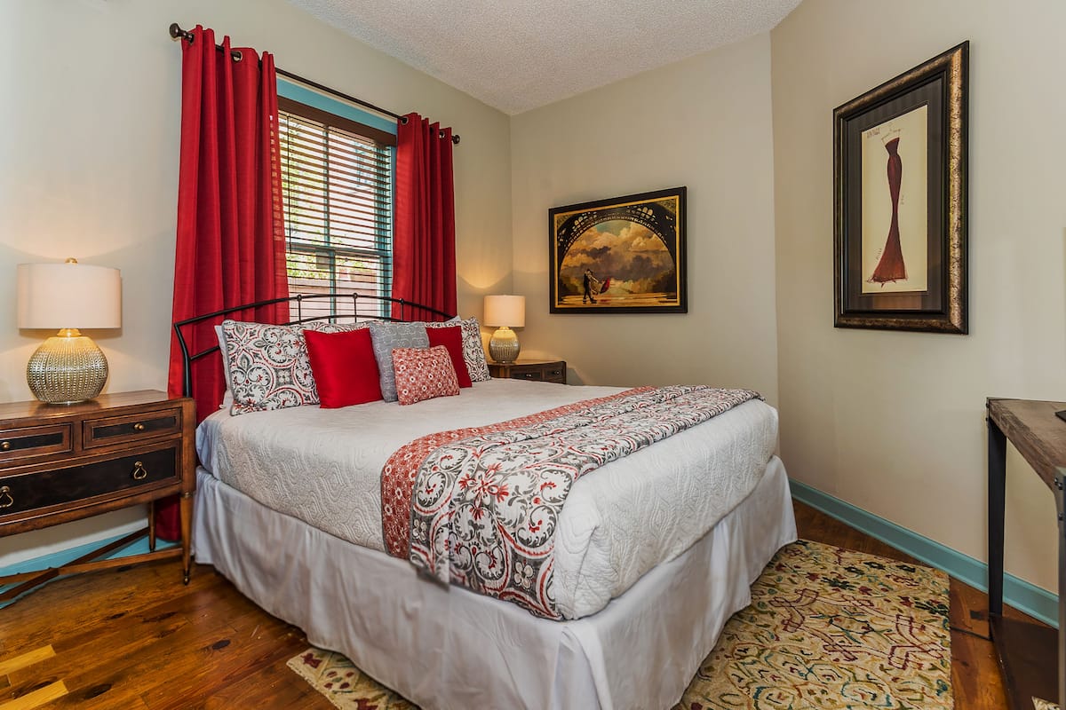 A neatly arranged bedroom with a bed, two bedside tables with lamps, a dresser, a window with red curtains, and a framed picture on the wall.
