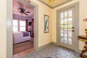 Una habitación de hotel con una cama, ventilador de techo, una puerta con ventanal y un cuadro en la pared.