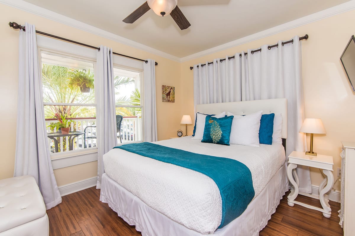 A bedroom with a large bed, a ceiling fan, a window with curtains, a small table with a lamp, and a potted plant on the windowsill.