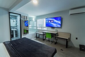 Una habitación de hotel moderna con una cama, un escritorio, una silla, un televisor y un baño visible a través de una puerta abierta.