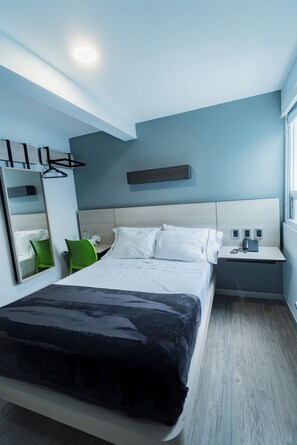 Una habitación de hotel moderna con una cama grande, una mesita de noche con teléfono y lámpara, un espejo y una silla verde.