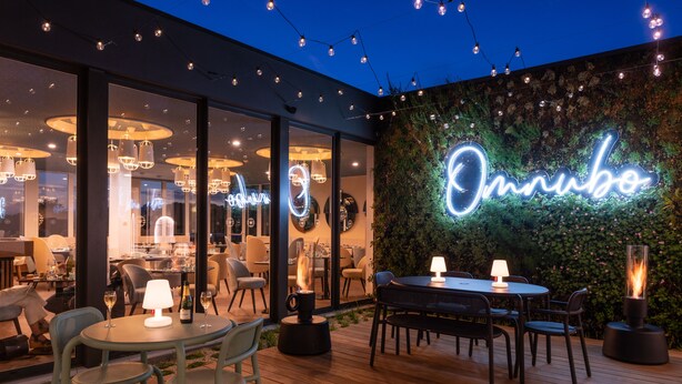 Un espace de restauration en plein air avec des tables et des chaises, un panneau lumineux au néon et des guirlandes lumineuses.